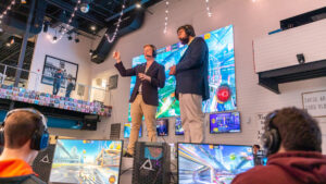 Courtesy Visit Myrtle Beach Two broadcasters stand on stage surrounded by competitors at collegiate esports competition, Boost at the Beach, held in Myrtle Beach, S.C.