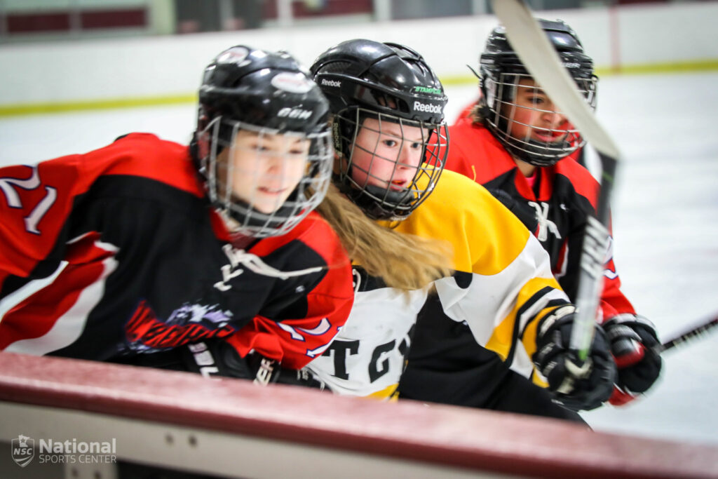 Girls Hockey players skate by the sideboards. 