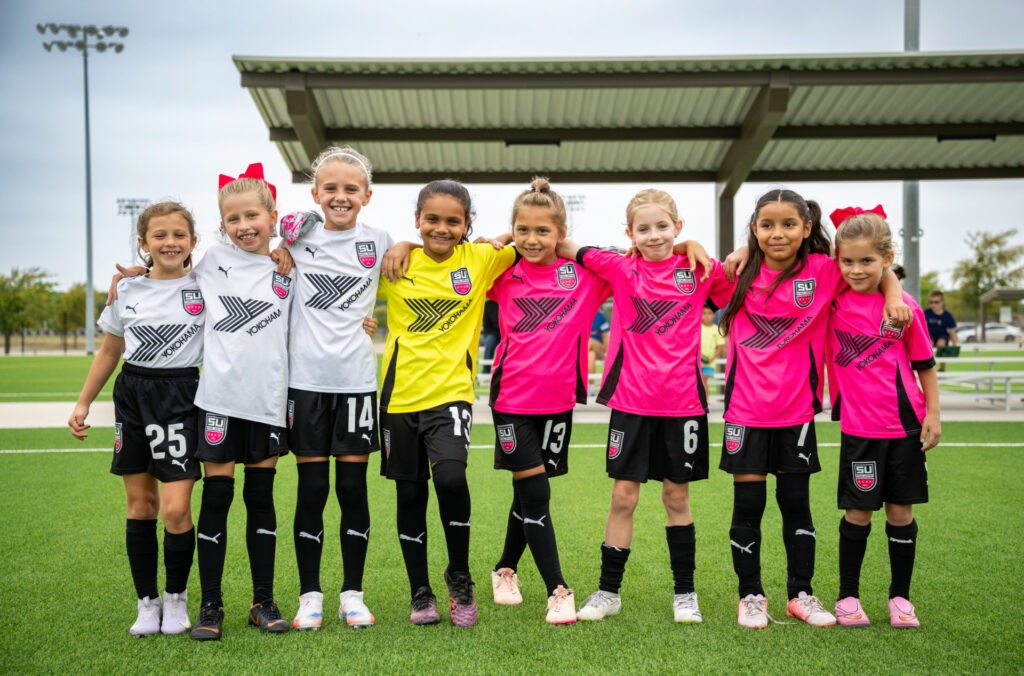 A group of girl soccer players standing side by side on a field.