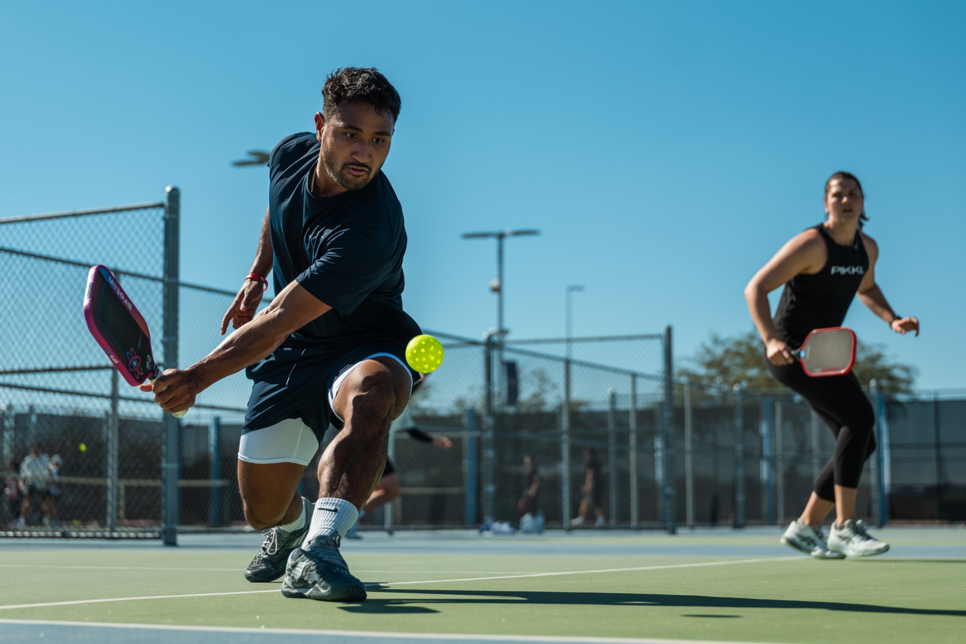 Pickleball players compete outdoors on a sunny court, with a man hitting the ball in the foreground and a woman playing behind him.