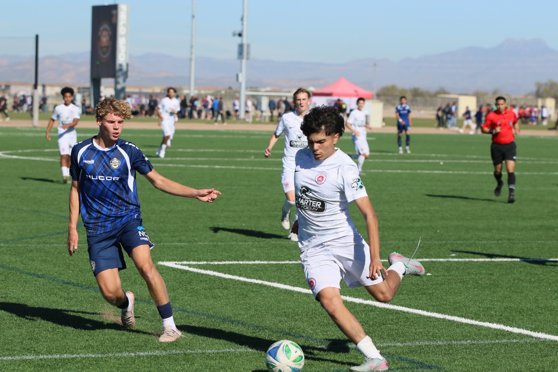Youth soccer players compete for possession of the ball on an outdoor artificial-turf field during a match, with teammates, spectators, and a referee in the background.