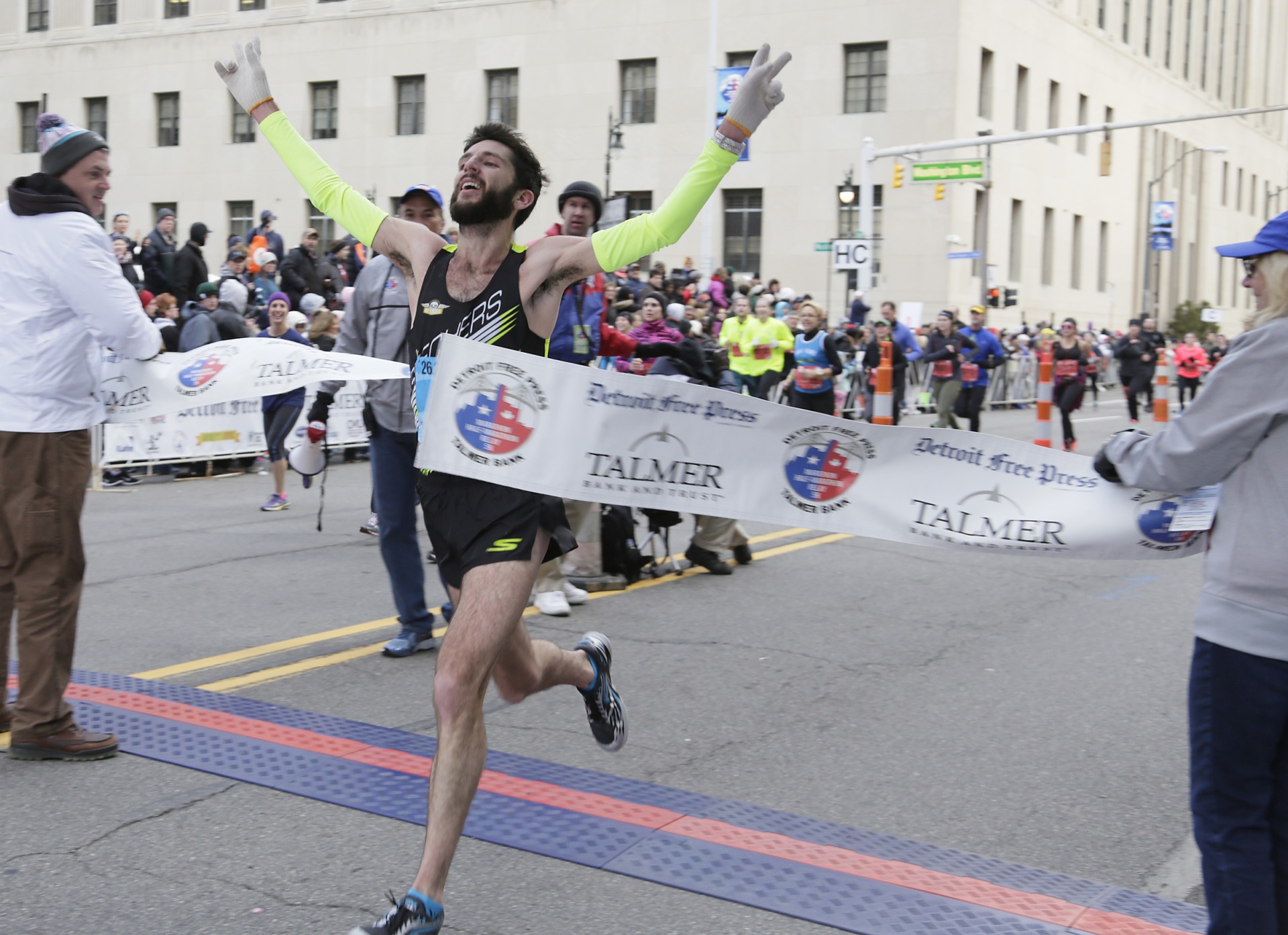 A male runner with his arms raised crosses the finish line during the Detroit Free Press Marathon as spectators cheer along a downtown Detroit street.